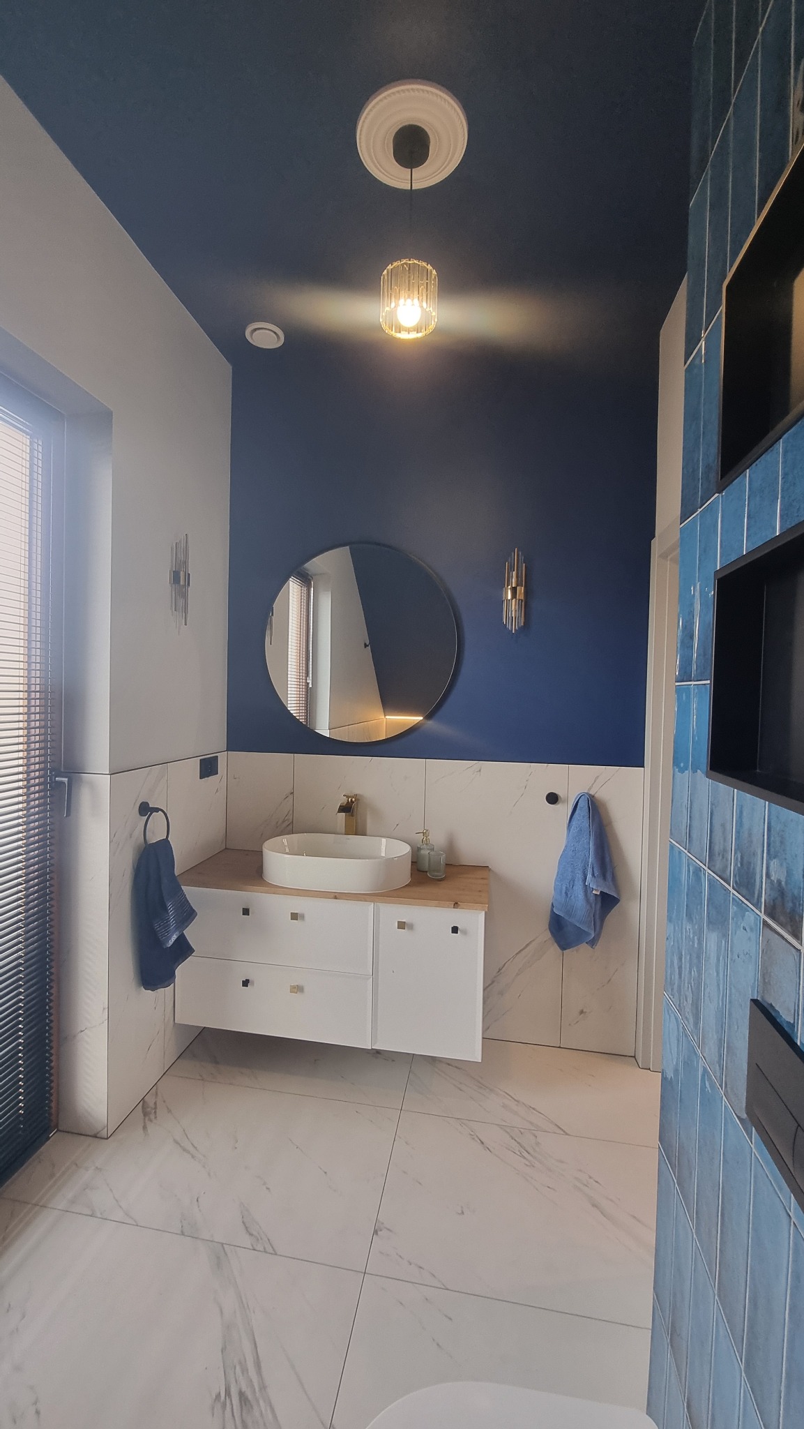 Nowoczesna łazienka z okrągłym lustrem, białą umywalką nablatową i niebieskimi akcentami kolorystycznymi na ścianach i płytkach. Widoczna dekoracyjna lampa sufitowa.