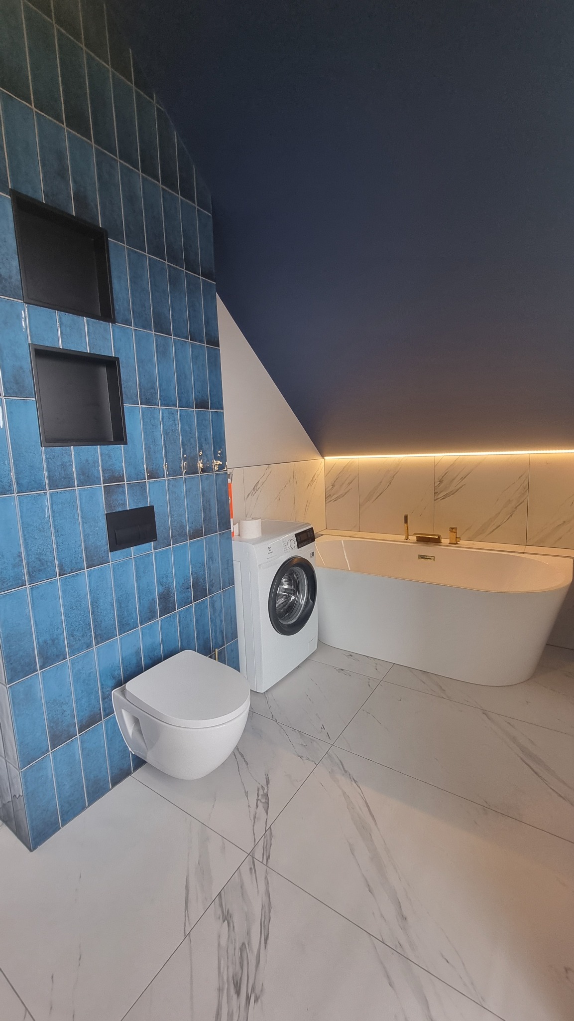 Niebieska łazienka na poddaszu z wanną, toaletą i pralką. Ściany wyłożone błyszczącymi, pionowymi płytkami. Podłoga z białego marmuru. Oświetlenie LED nad wanną.