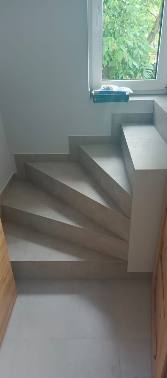 Wykończone schody wewnętrzne z szarymi płytkami, widok z góry. Na parapecie okna leżą narzędzia. Fragment drewnianych drzwi po prawej stronie kadru.