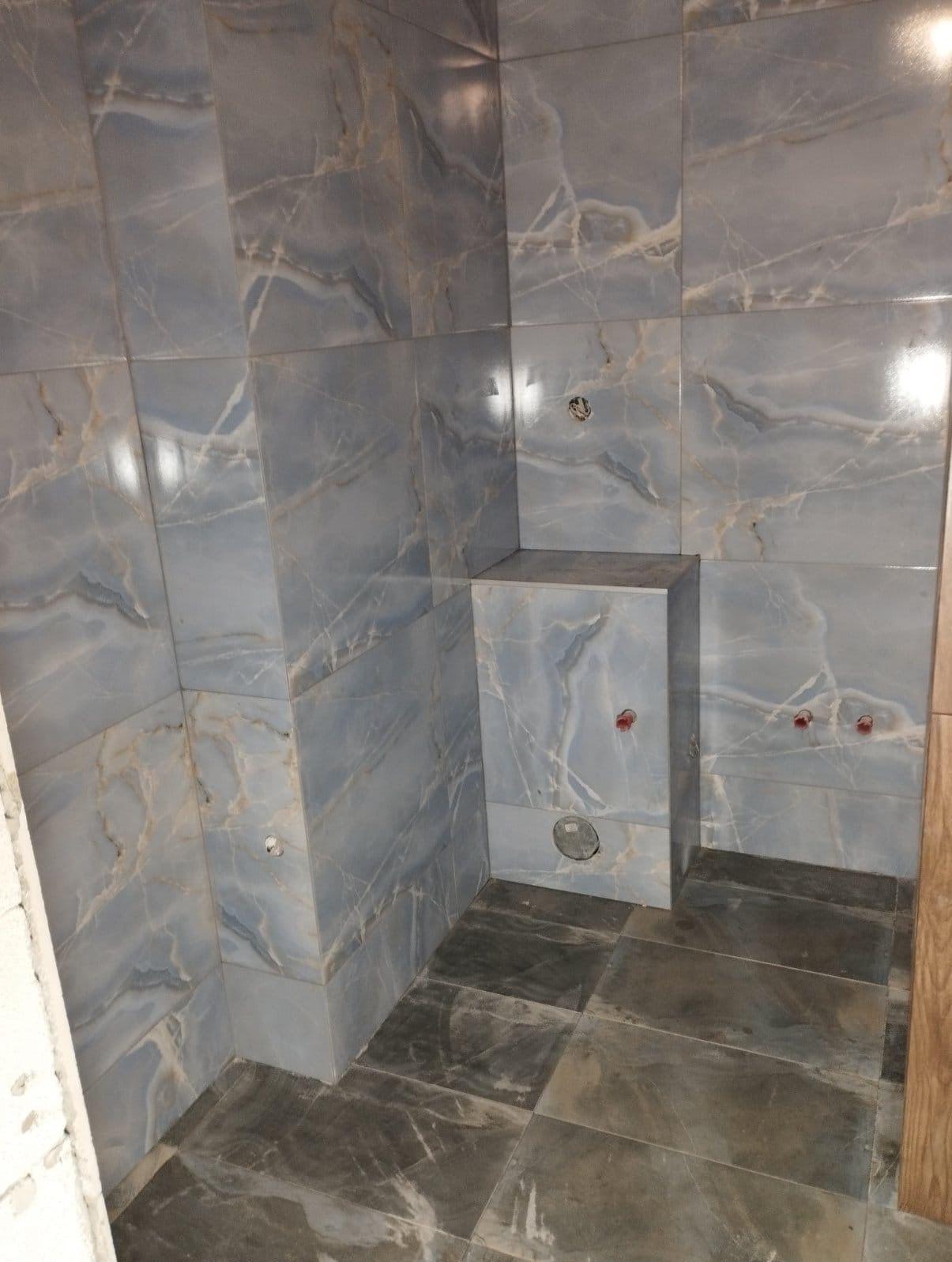 Wykończona łazienka z szaro-niebieskimi płytkami imitującymi marmur na ścianach i podłodze. Widoczne przyłącza wodne i zabudowa geberitu.