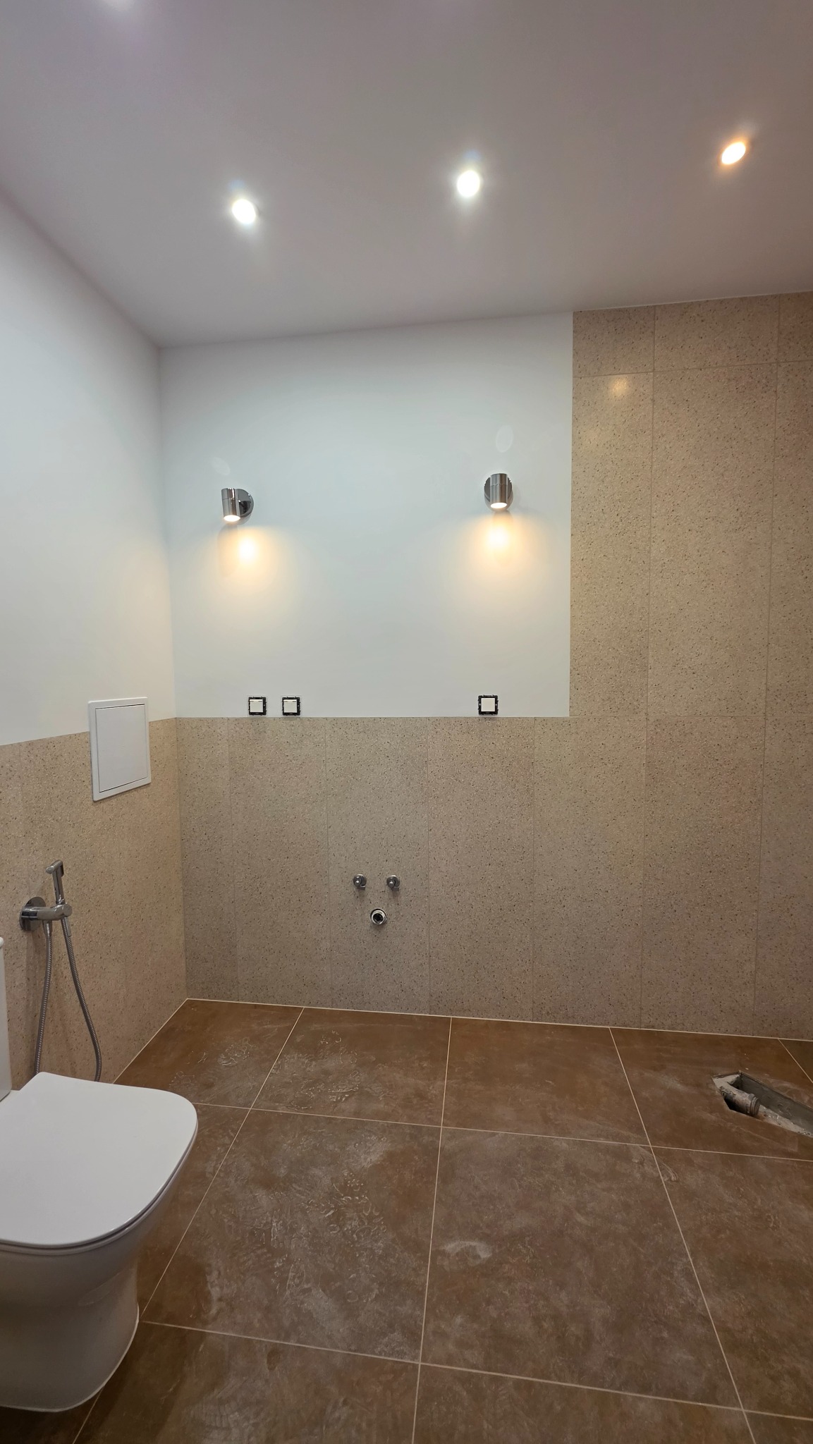 Wykończona łazienka w Bydgoszczy z beżowymi płytkami na ścianach i brązowymi na podłodze. Widoczne chromowane lampy i armatura. Częściowo widoczna toaleta.