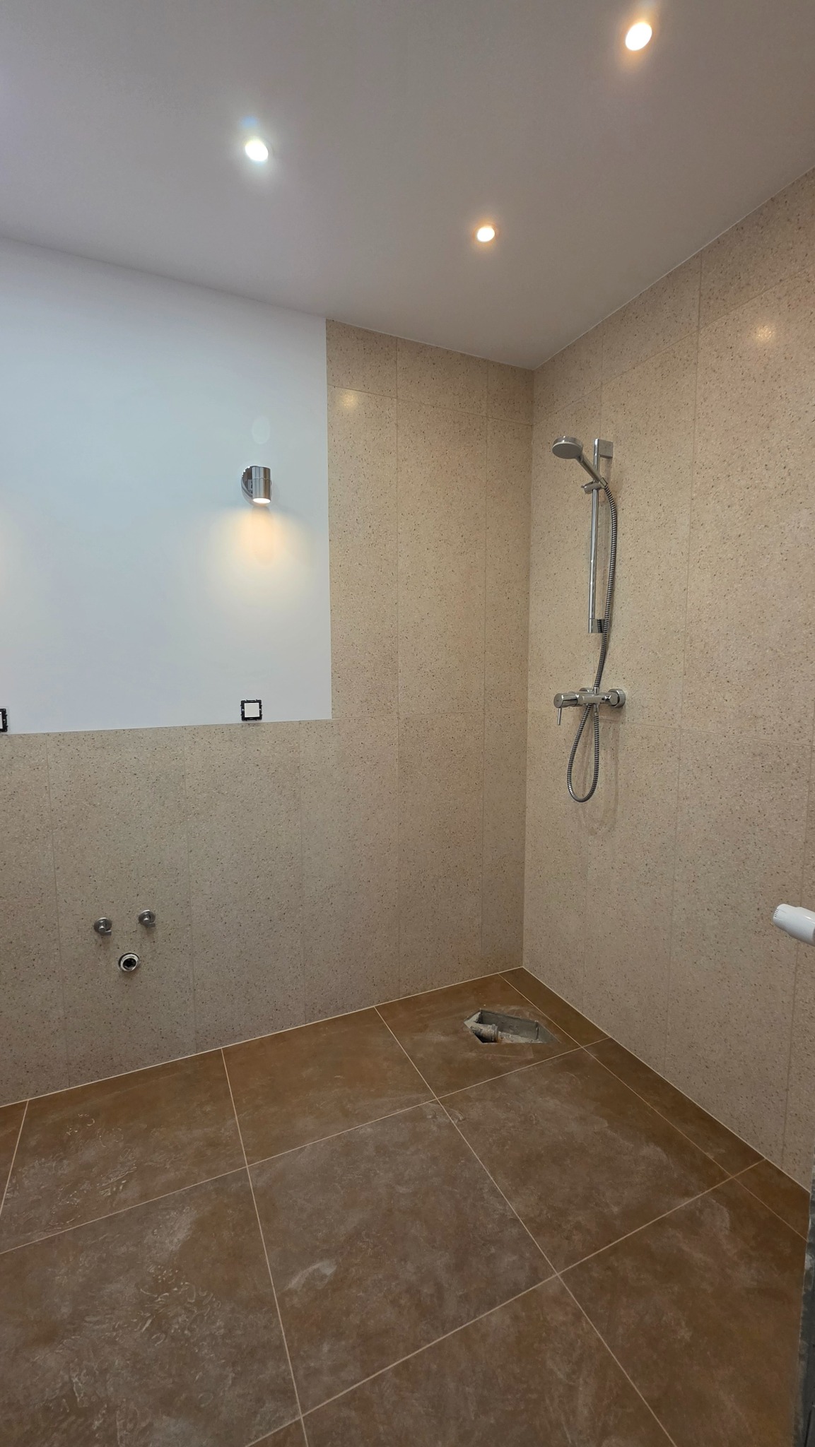 Wykończona łazienka z beżowymi płytkami na ścianach i brązowymi na podłodze. Widoczna armatura prysznicowa i przygotowane punkty instalacyjne.