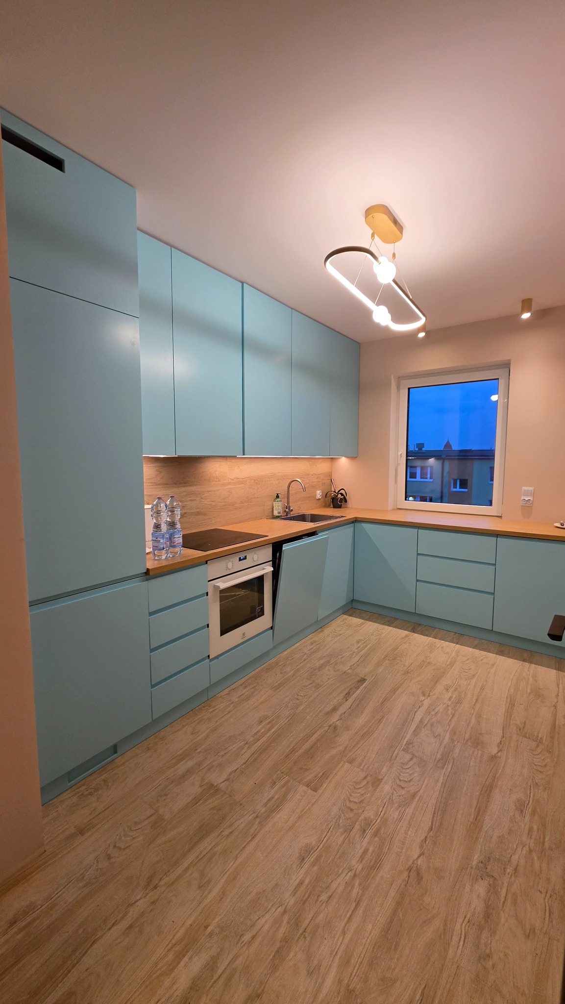 Błękitna kuchnia z drewnianym blatem i podłogą. Nowoczesne oświetlenie sufitowe. Widok na budynek z okna. Dwie butelki wody na blacie.