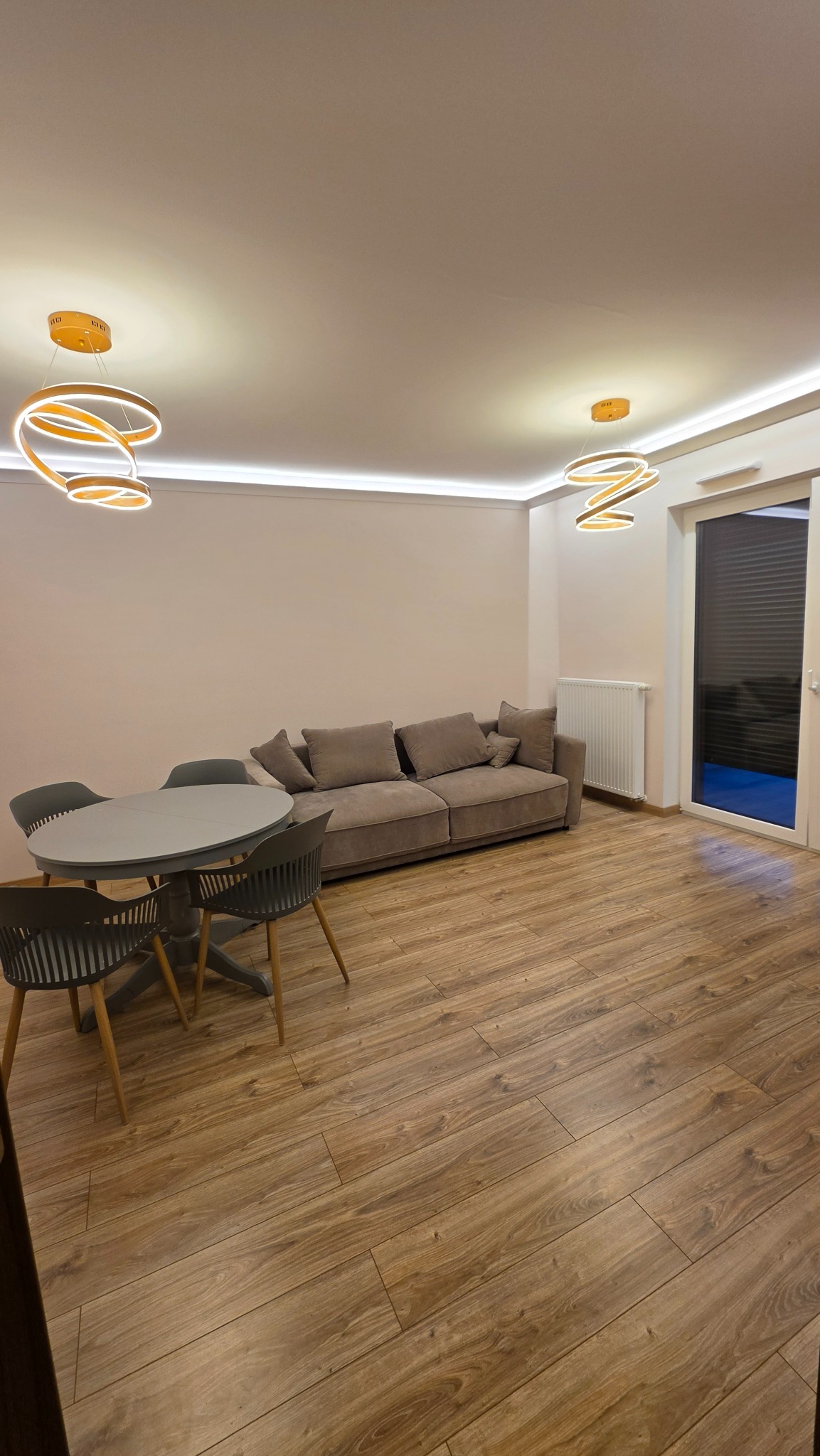 Jasne wnętrze pokoju z drewnianą podłogą, szarą sofą i okrągłym stołem. Dwa nowoczesne, okrągłe żyrandole z ciepłym światłem. Widoczne listwy LED pod sufitem.