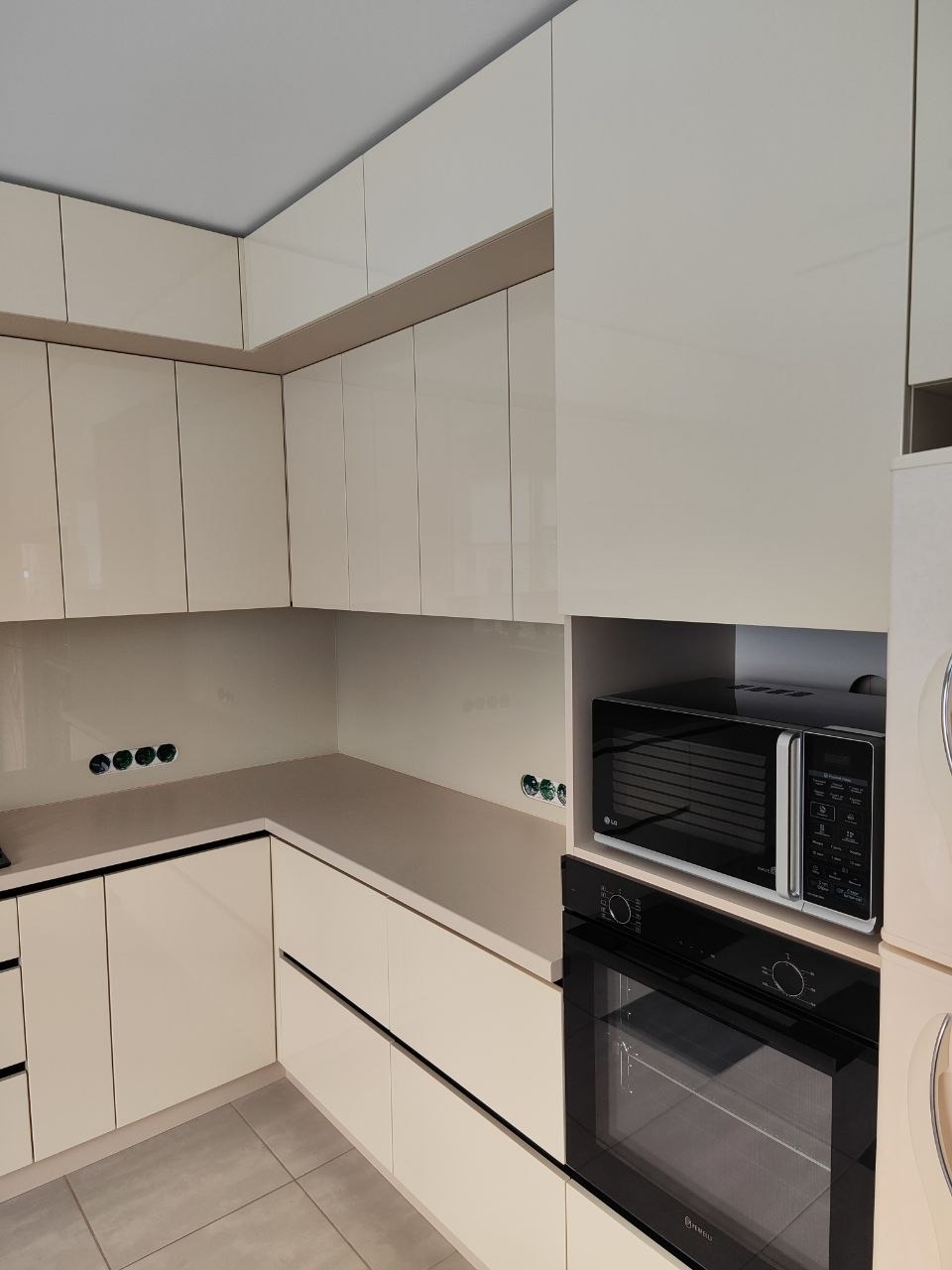 Beżowa, narożna zabudowa kuchenna z piekarnikiem i mikrofalówką w nowoczesnym stylu. Widoczne gniazdka elektryczne w ścianie nad blatem.