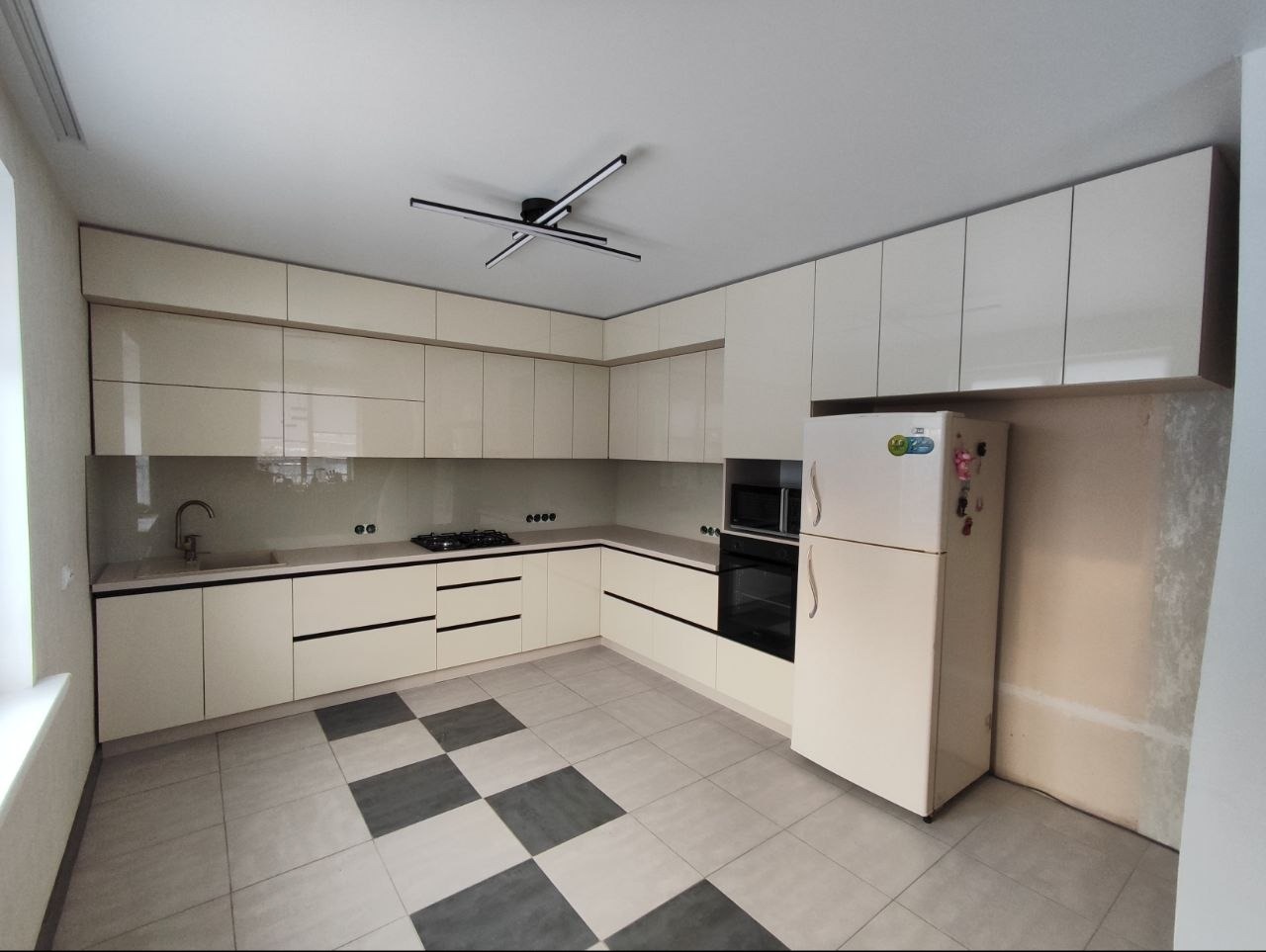 Jasna kuchnia z błyszczącymi frontami, czarnymi uchwytami i szachownicą na podłodze. Widoczny fragment ściany przed wykończeniem obok lodówki.