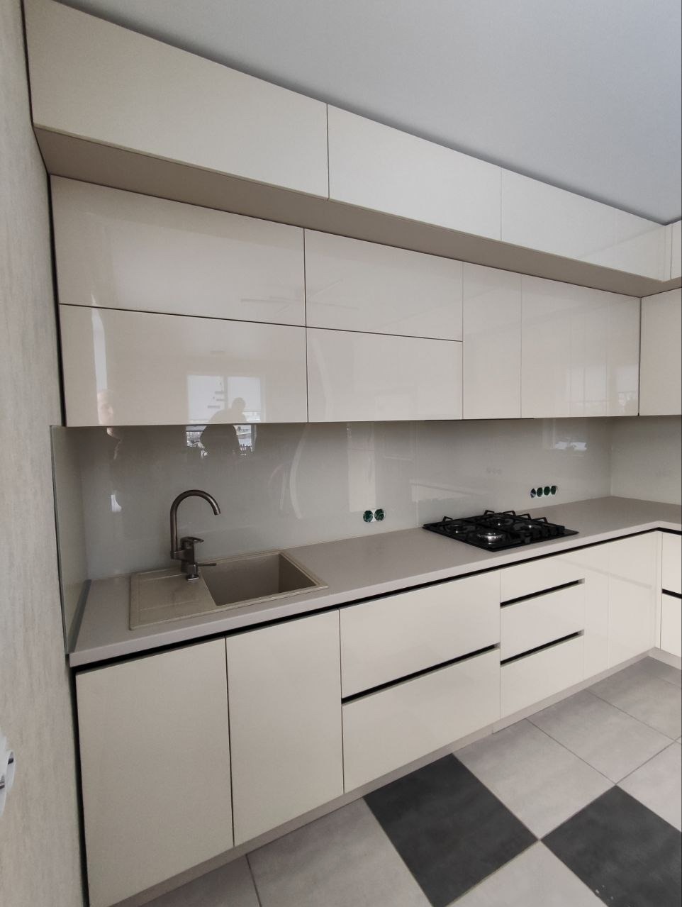 Nowoczesna, jasna kuchnia z błyszczącymi frontami szafek, szarym blatem i płytkami podłogowymi w szachownicę. Minimalistyczny design z widoczną płytą grzewczą i zlewem.