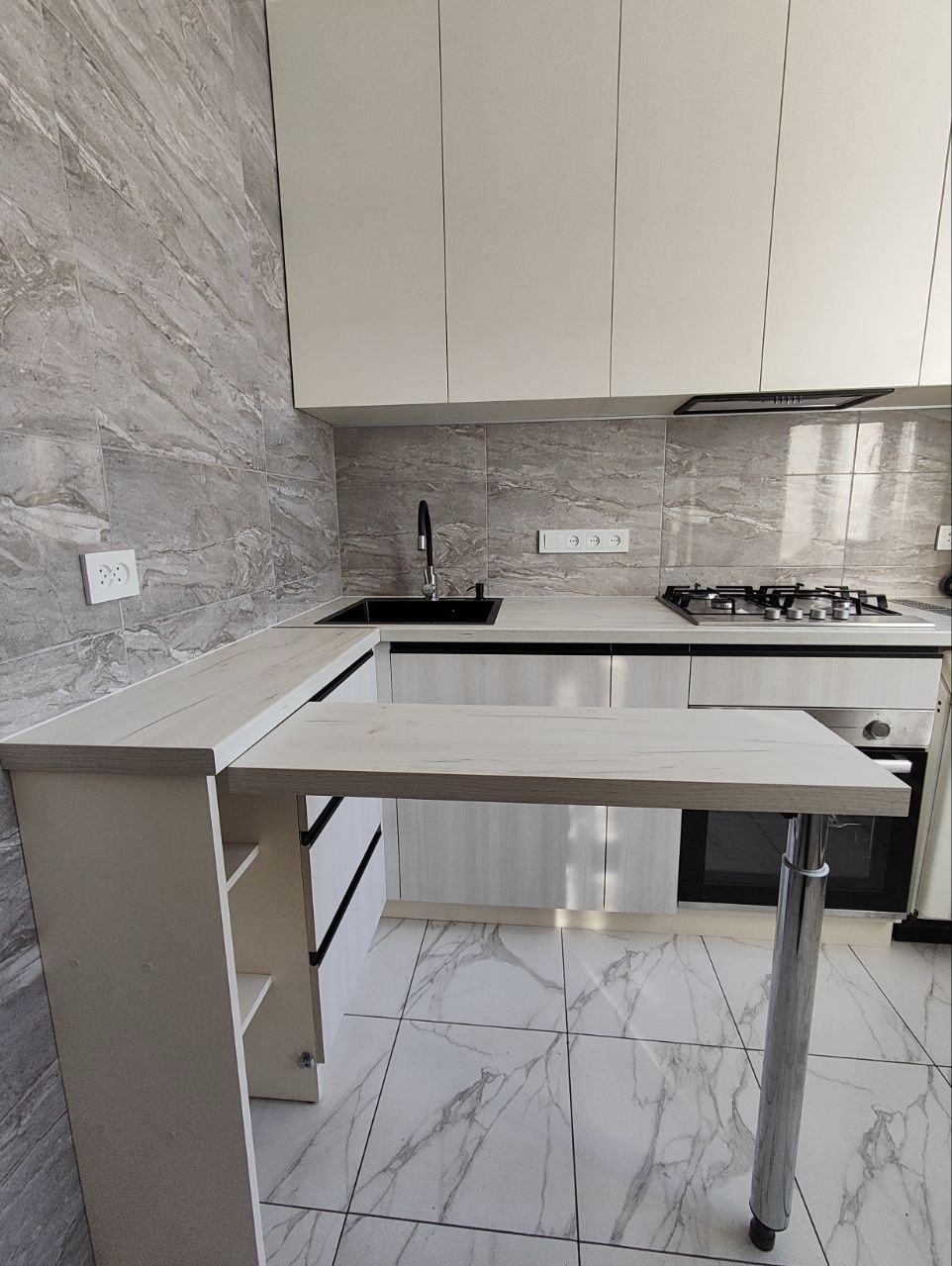 Jasna kuchnia z blatem barowym na chromowanej nodze, marmurowe płytki na ścianie i podłodze. Nowoczesne wykończenie, minimalistyczny design.