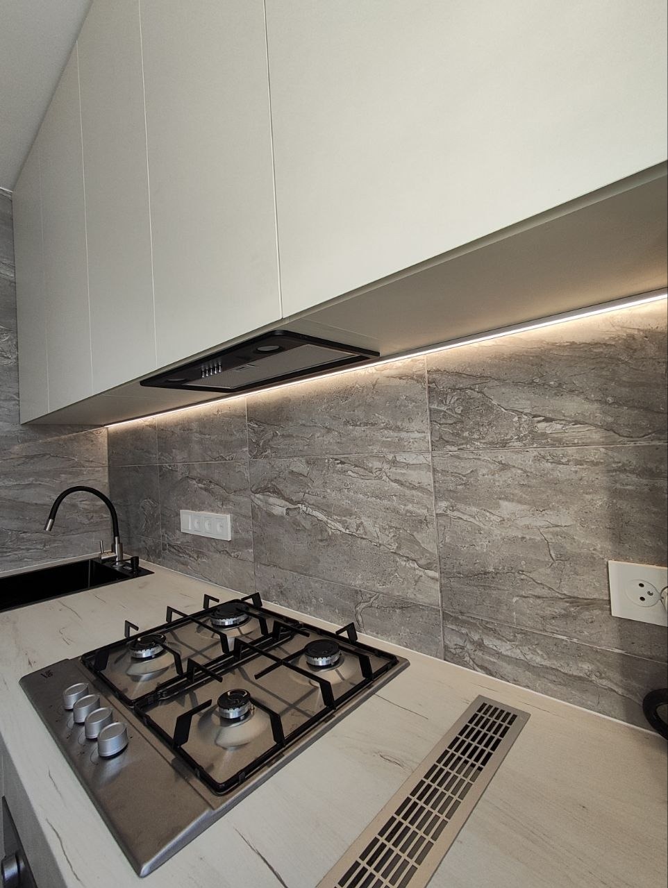 Nowoczesna kuchnia z szarymi płytkami imitującymi marmur, białymi szafkami i oświetleniem LED pod szafkami. Widoczna płyta gazowa i czarny zlew.