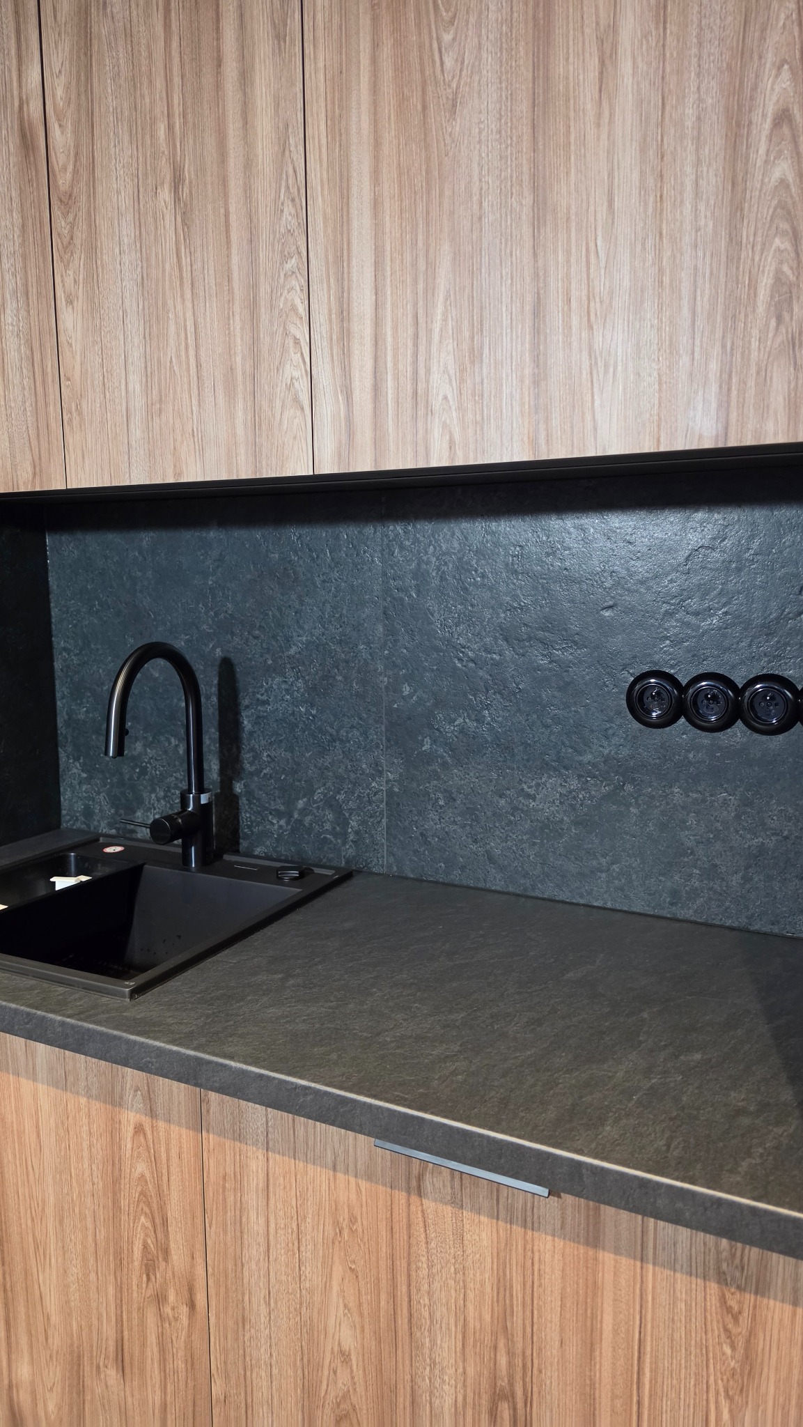 Nowoczesna kuchnia: drewniane szafki, czarny zlew i blat z kamienną fakturą. Uwagę przykuwają retro gniazdka na tle ściany o surowym wykończeniu.