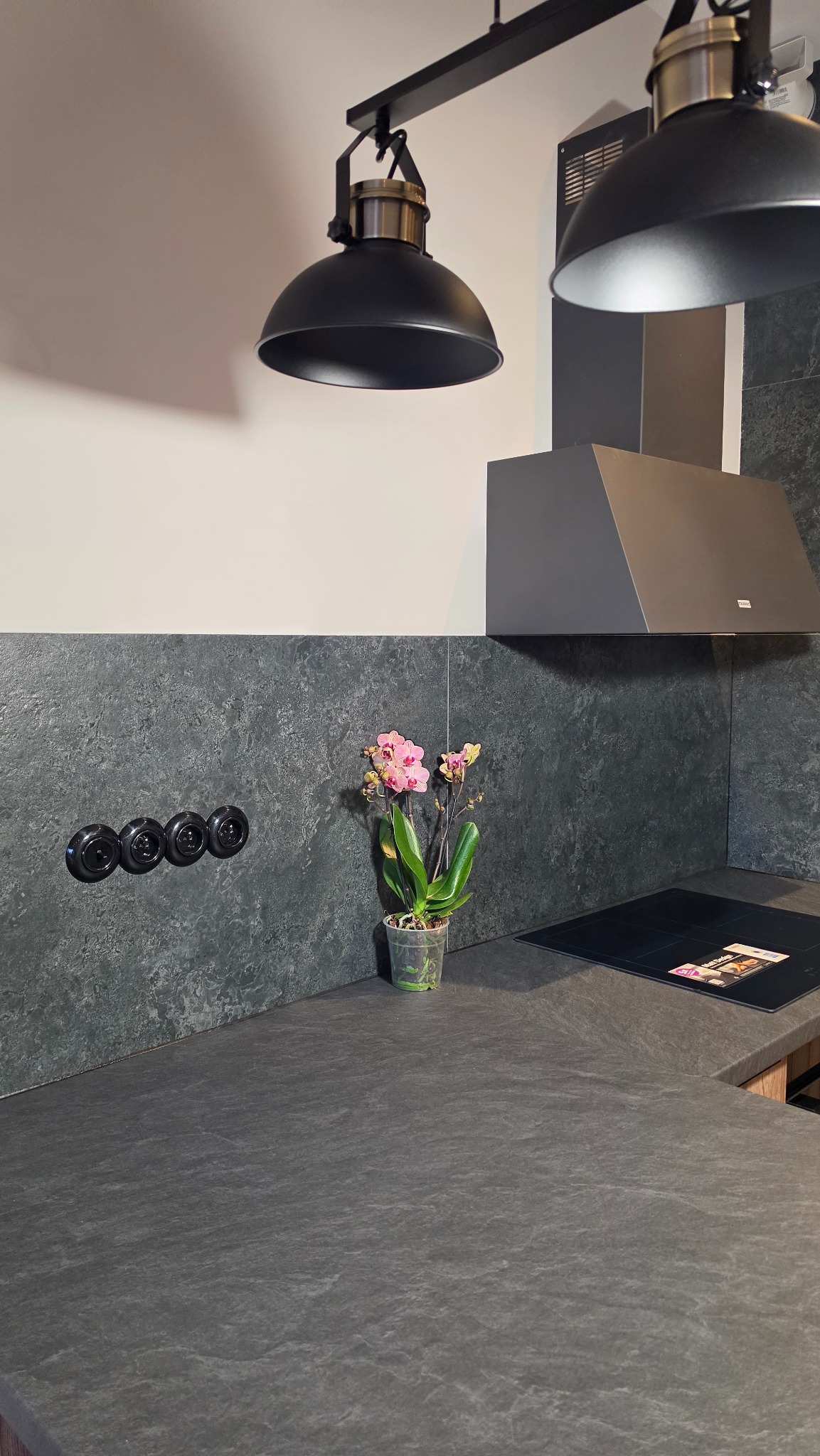 Nowoczesna kuchnia z blatem z szarego kamienia, czarną płytą indukcyjną, okapem i oświetleniem. Na blacie stoi storczyk w przezroczystej doniczce. Na ścianie włączniki.