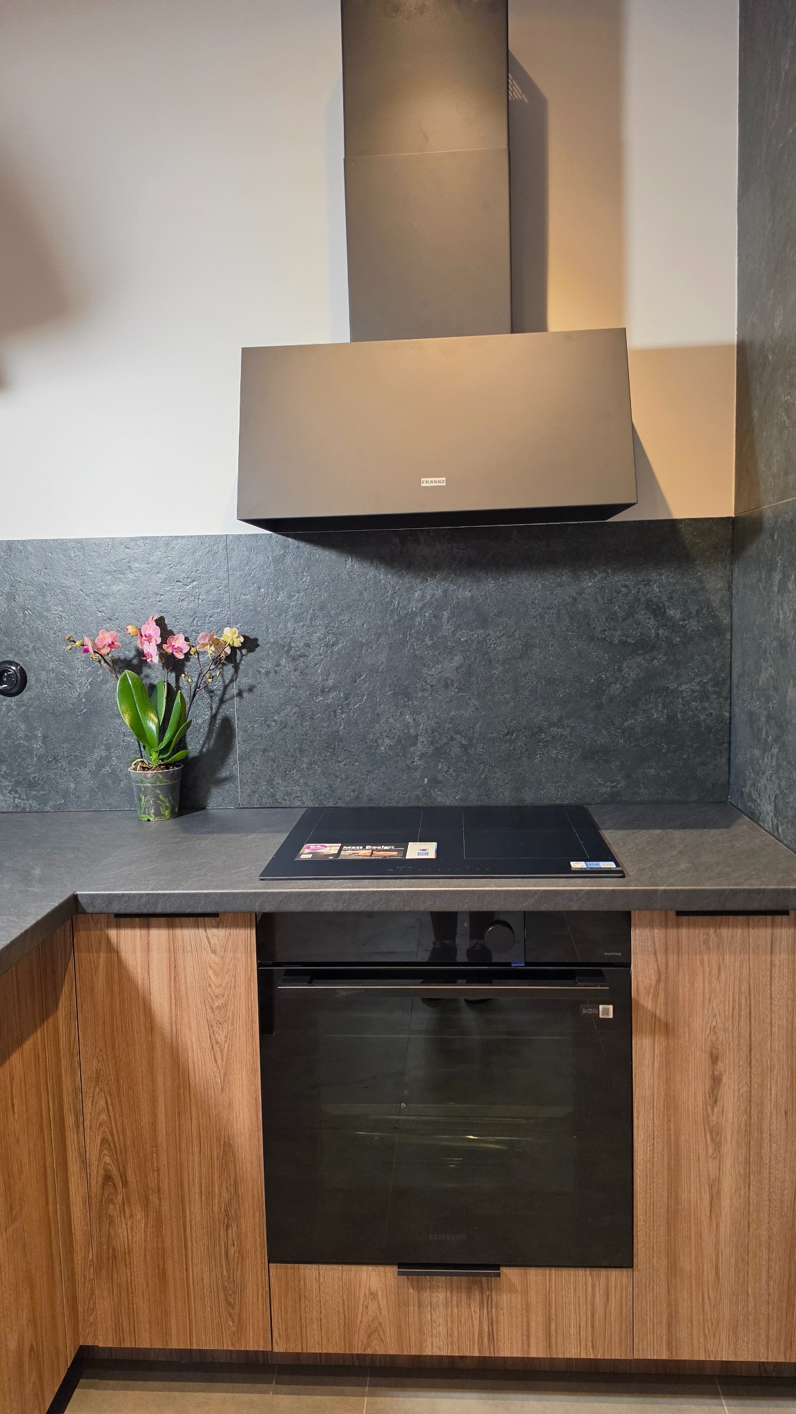 Nowoczesna kuchnia z drewnianymi szafkami, czarnym blatem i płytą indukcyjną. Nad płytą znajduje się czarny okap marki Franke. Na blacie stoi wazon z kwiatami.