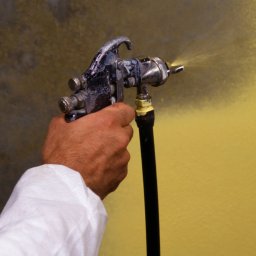 Dr Myjka - Malowanie natryskowe ściany na żółto. W dłoni pistolet lakierniczy, widoczny wąż i fragment ściany z efektem rozpylonej farby. Osoba w kombinezonie ochronnym.