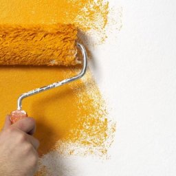 Dr Myjka - Malowanie ściany wałkiem na złoto. Dłoń trzyma narzędzie, widoczny fragment pomalowanej i surowej ściany. Tekstura farby i wałka.