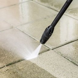 Dr Myjka - Czyszczenie kostki brukowej myjką ciśnieniową. Strumień wody usuwa zabrudzenia, ukazując czystą powierzchnię. Widoczne fugi i dysza myjki.