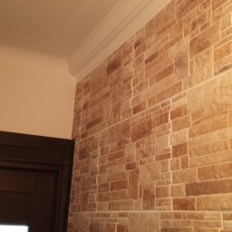 Dr Myjka - Dekoracyjna ściana z imitacji kamienia w odcieniach beżu i brązu, wykończona białym, ozdobnym gzymsem przy suficie, widoczny fragment ciemnych drzwi z poziomymi przeszkleniami.