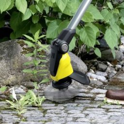 Dr Myjka - Elektryczna szczotka do usuwania chwastów Kärcher w akcji, czyszcząca kamienną nawierzchnię. W tle widoczne zielone liście krzewów i fragment buta.