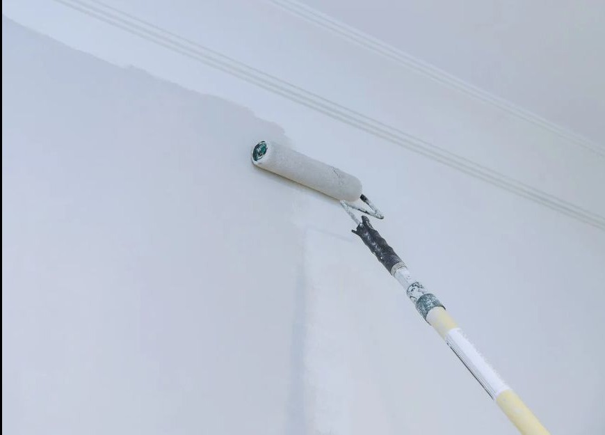 Malowanie ściany wałkiem teleskopowym na biało. Widoczna świeżo pomalowana powierzchnia i fragment listwy przysufitowej. Praca w toku.