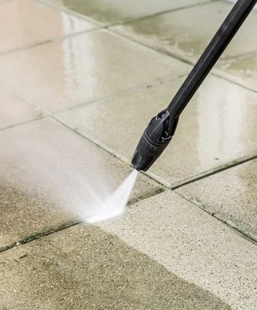 Czyszczenie kostki brukowej myjką ciśnieniową. Strumień wody usuwa zabrudzenia, ukazując czystą powierzchnię. Widoczne fugi i dysza myjki.