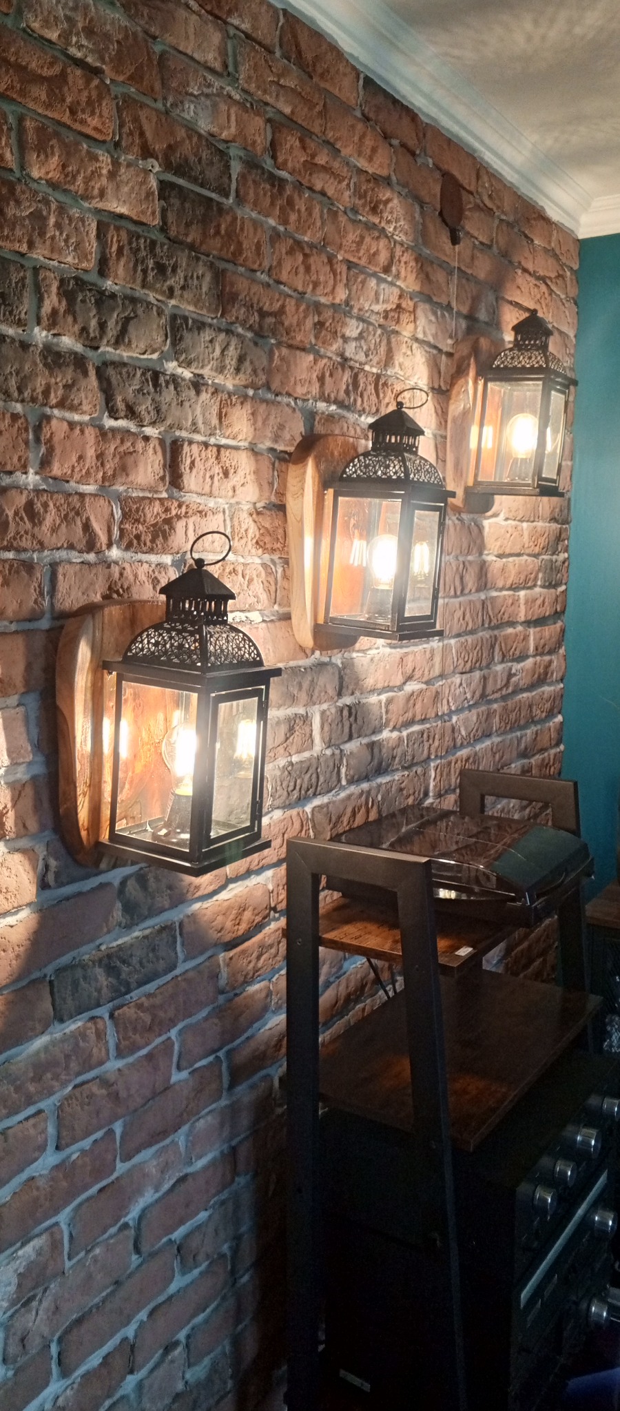 Dekoracyjna ściana z cegły z trzema lampionami na drewnianych podstawkach, oświetlającymi wnętrze. Widoczny fragment regału.