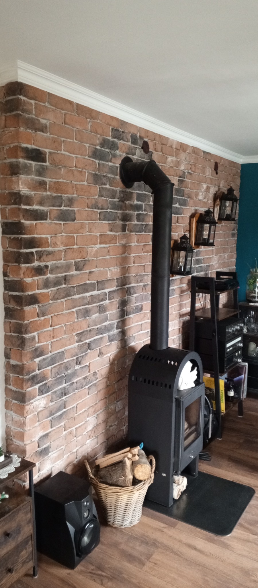 Ceglana ściana z kominkiem i koszem drewna, wzbogacona o biały sufitowy gzyms i wiszące lampiony, na drewnianej podłodze. Akcenty dekoracyjne w tle.