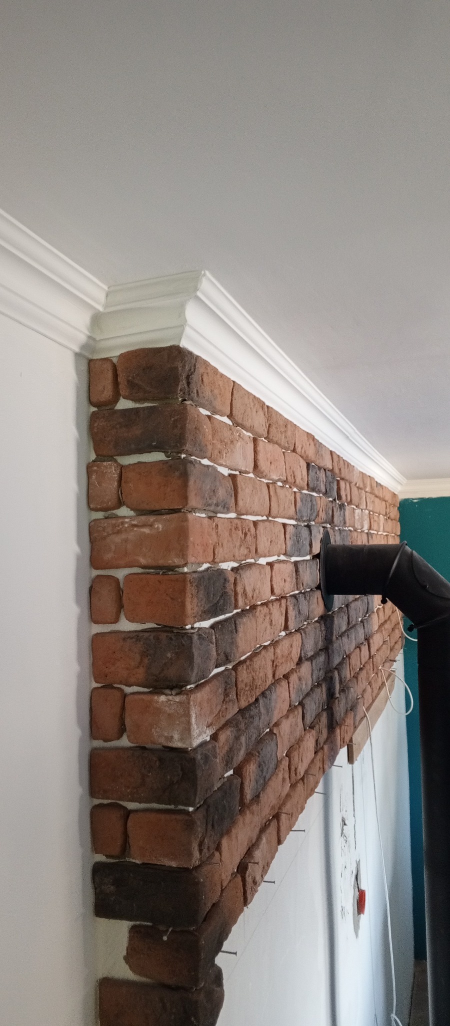 Ściana wykończona dekoracyjną cegłą w odcieniach brązu i czerni, z białym ozdobnym gzymsem pod sufitem i fragmentem czarnego komina. Widok z dołu, podkreślający fakturę.