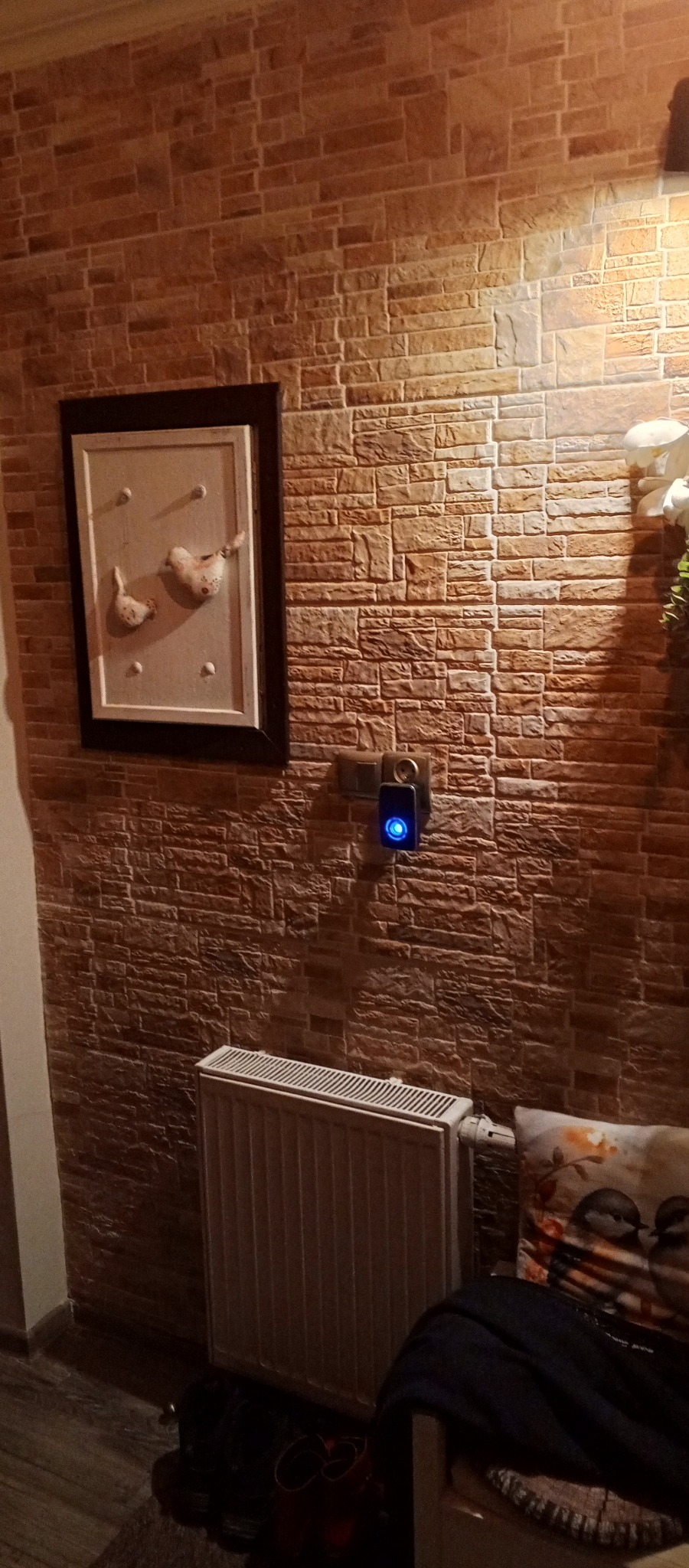 Ściana z imitacji cegły, ozdobiona obrazkiem z ptakami i nowoczesnym dzwonkiem. Grzejnik i dekoracyjna poduszka dopełniają wnętrze.