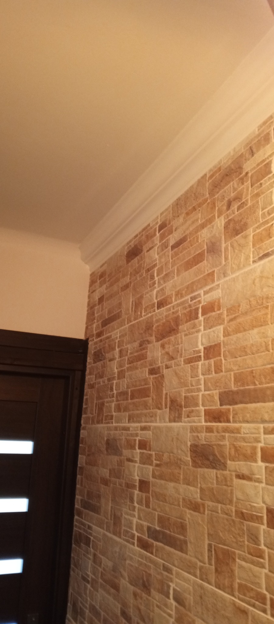 Dekoracyjna ściana z imitacji kamienia w odcieniach beżu i brązu, wykończona białym, ozdobnym gzymsem przy suficie, widoczny fragment ciemnych drzwi z poziomymi przeszkleniami.
