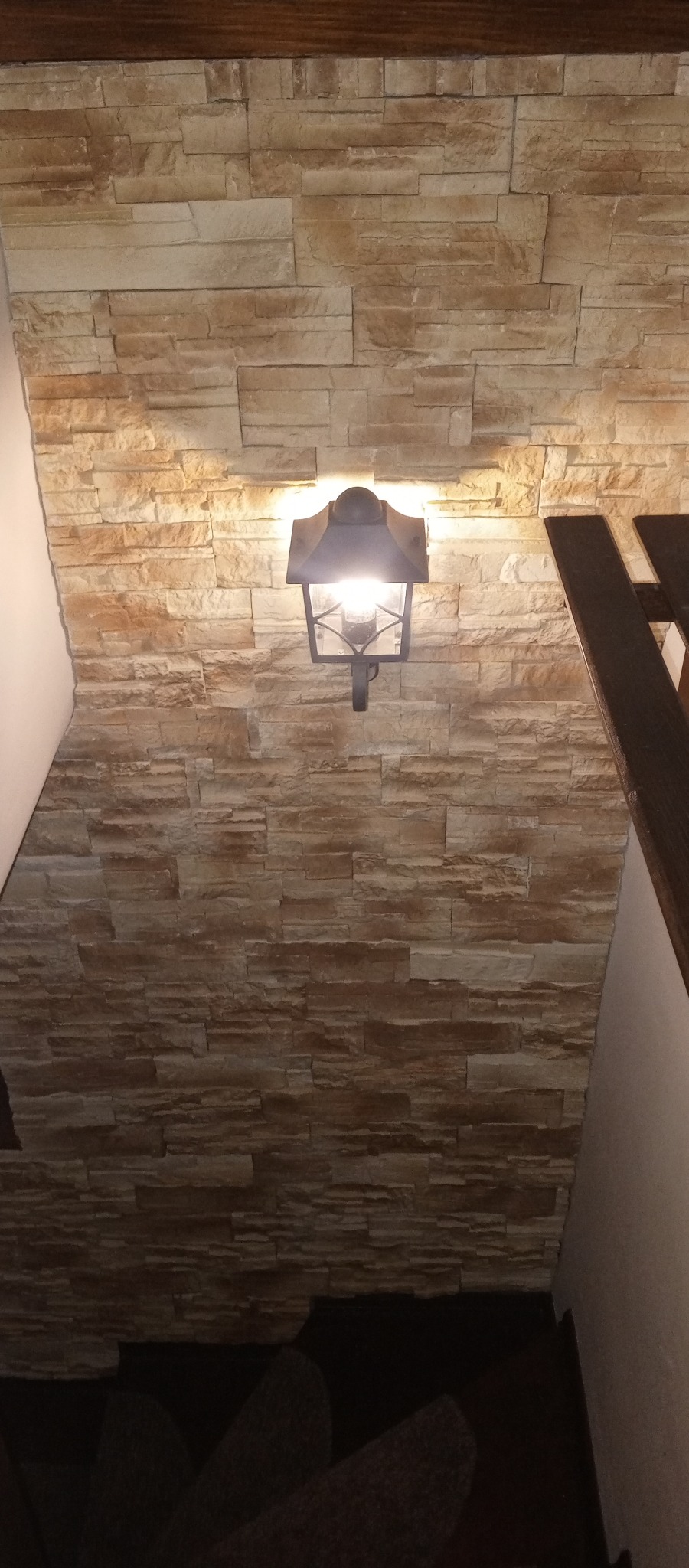 Ściana wyłożona dekoracyjną, beżową cegłą, oświetlona przez metalowy kinkiet w stylu retro. Fragment schodów widoczny po prawej stronie kadru.