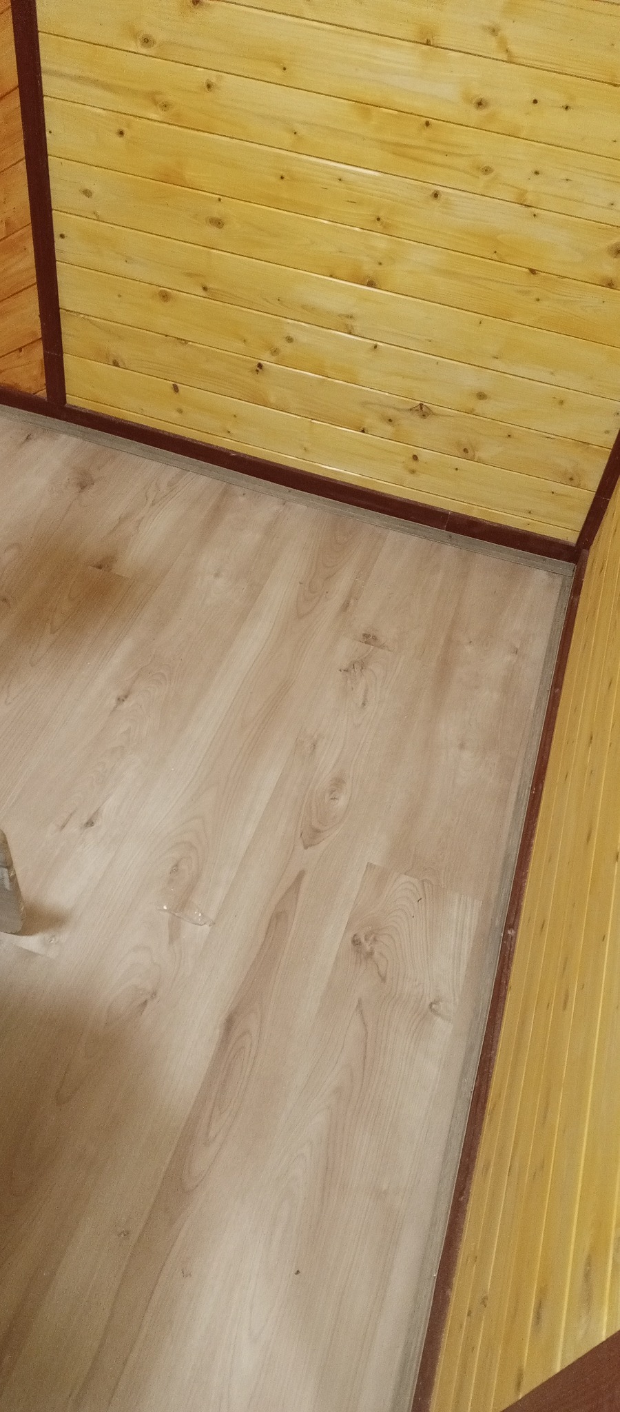 Jasne panele podłogowe ułożone w rogu pomieszczenia z boazerią, wykończone brązowymi listwami. Widok z góry, ukośny kadr.