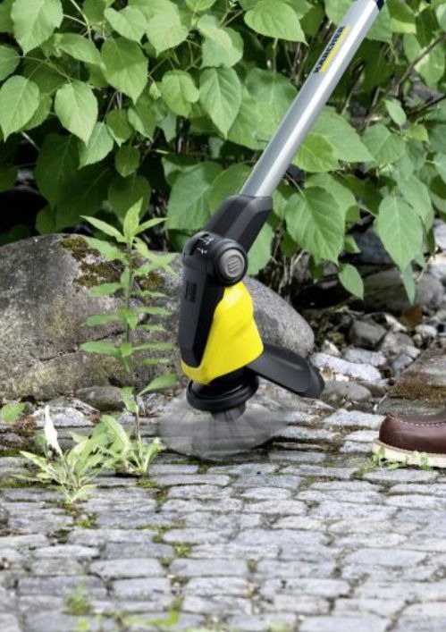Elektryczna szczotka do usuwania chwastów Kärcher w akcji, czyszcząca kamienną nawierzchnię. W tle widoczne zielone liście krzewów i fragment buta.