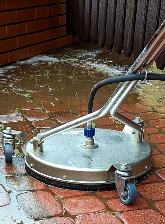 Czyszczenie kostki brukowej myjką ciśnieniową z okrągłą szczotką, widoczne mokre kostki i fragment muru. Profesjonalne narzędzie w trakcie pracy.