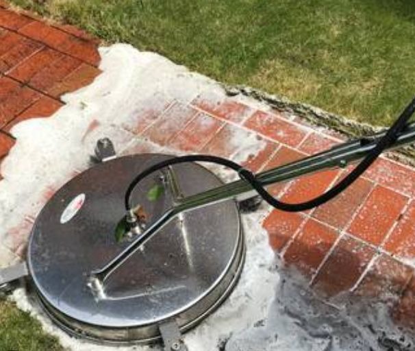 Czyszczenie kostki brukowej myjką ciśnieniową z okrągłą szczotką. Piana czyszcząca na czerwonej kostce brukowej obok trawnika. Widoczne narzędzie pracy.