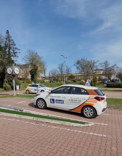 Plac manewrowy szkoły jazdy L-mot z dwoma samochodami Hyundai i20 w barwach firmy. Zegar na słupie odmierzający czas kursantom na tle zieleni i błękitnego nieba.