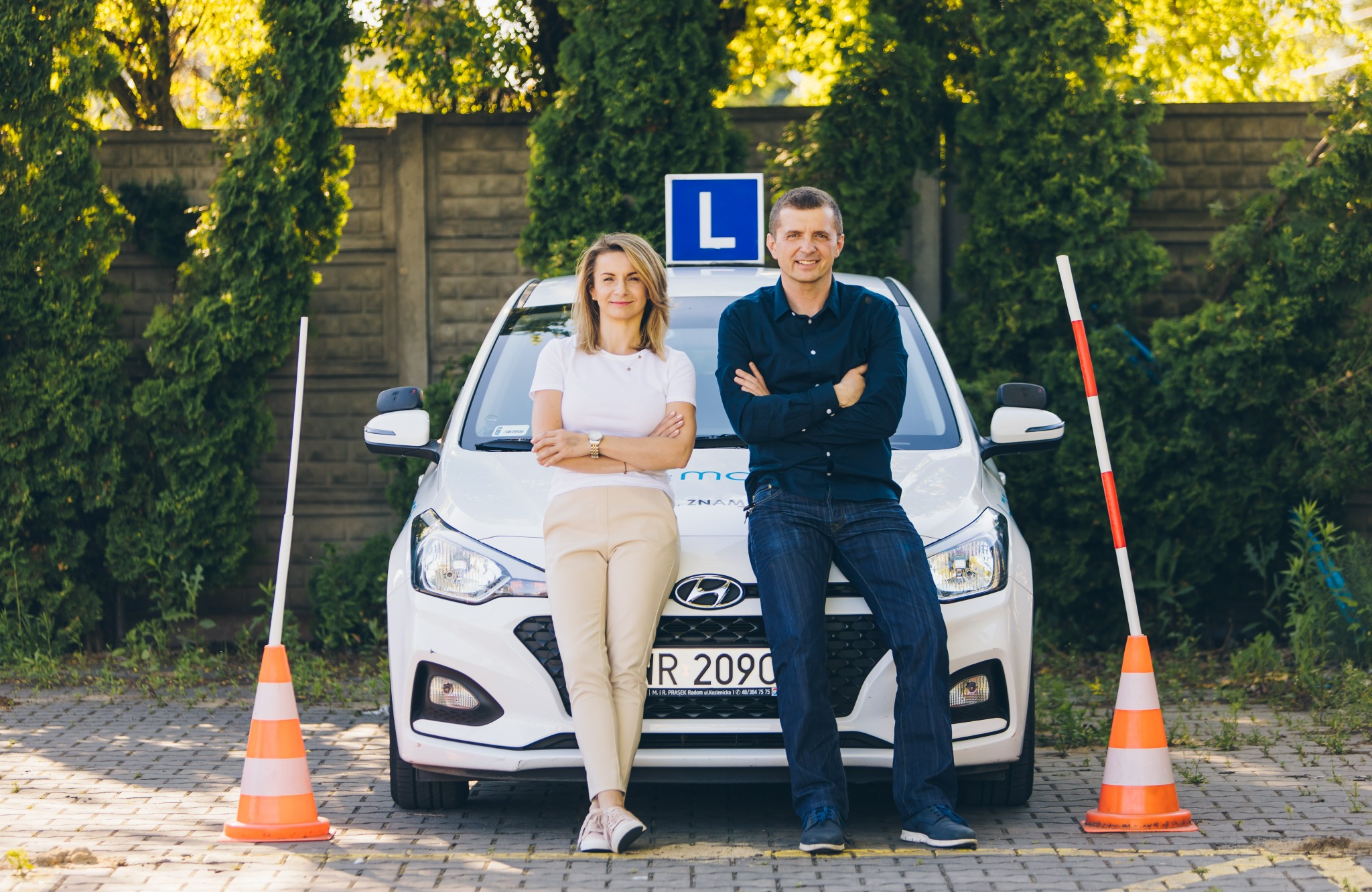 Instruktorzy szkoły jazdy pozują przy białym samochodzie z literą 'L', otoczonym pachołkami i tyczkami na tle zielonych krzewów i betonowego muru.