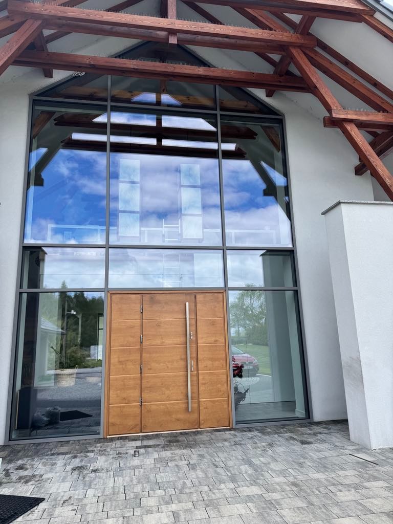 Duże okna z drewnianą stolarką odbijają błękitne niebo nad nowoczesnym domem z drewnianą konstrukcją dachu i szarą kostką brukową.