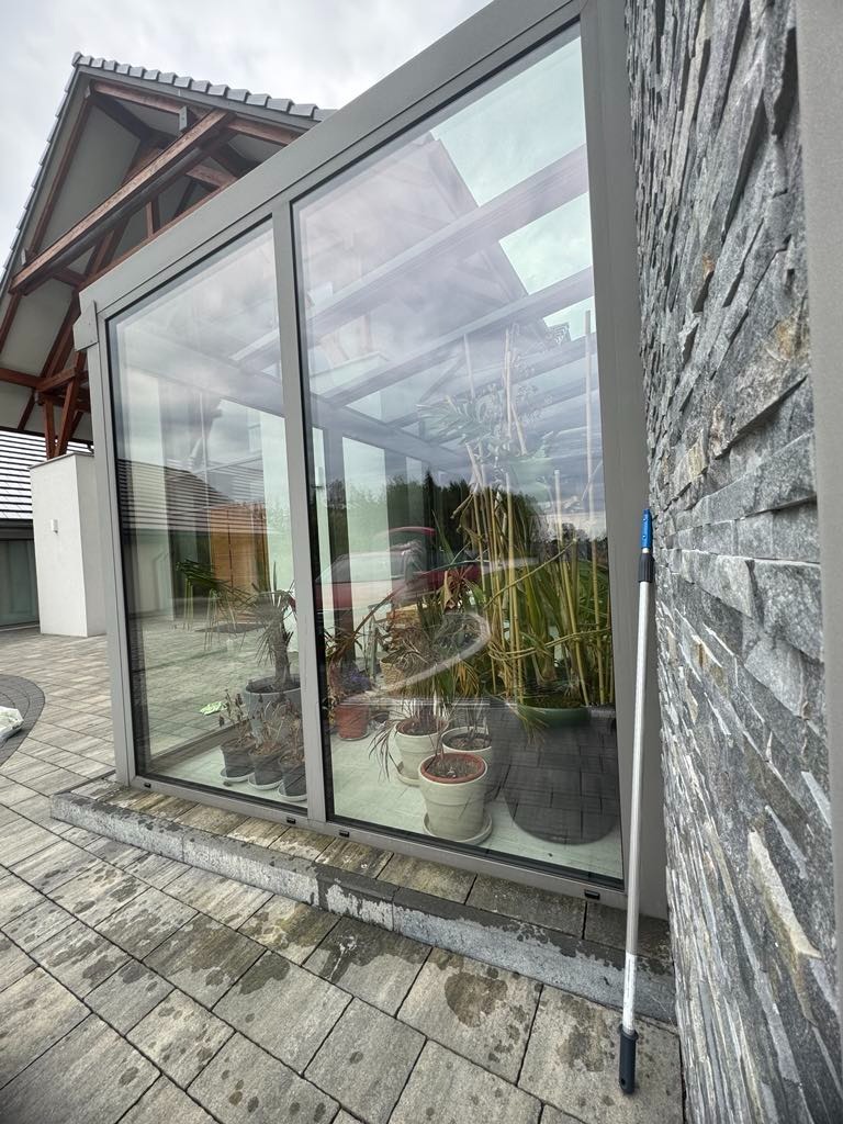 Szklana oranżeria z roślinami w donicach, widok z zewnątrz. Opierający się o ścianę kij teleskopowy do mycia okien. Kamienna elewacja i kostka brukowa.