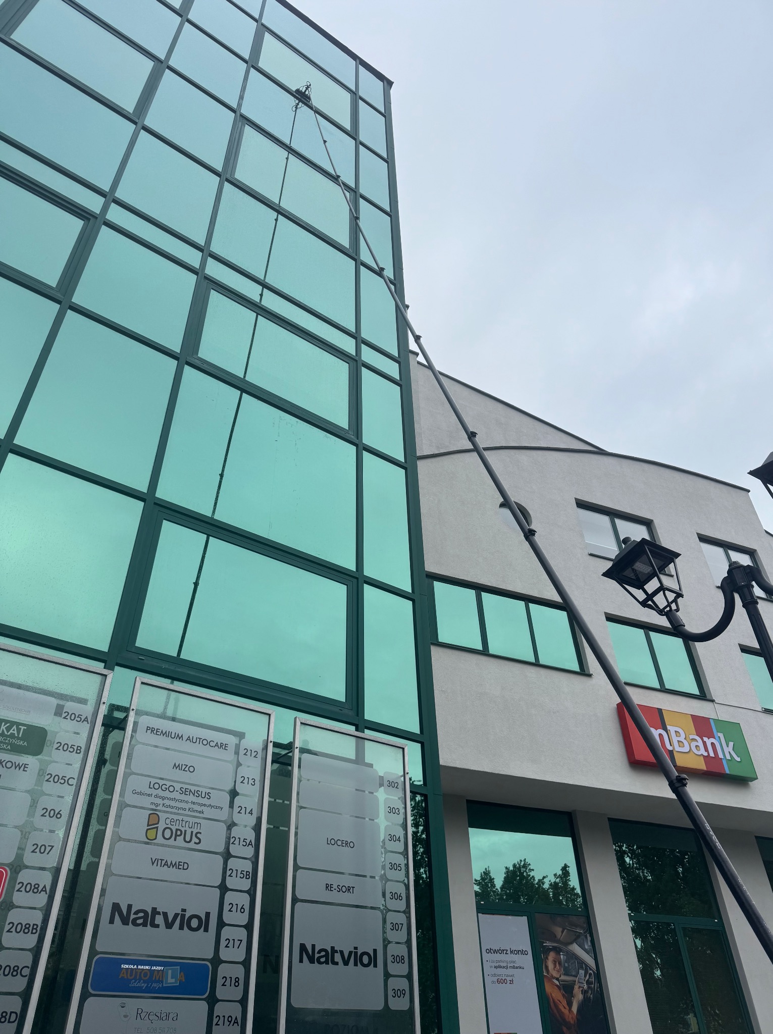Mycie okien na wysokości w biurowcu z zielonymi szybami. Długa lanca z szczotką sięga wysoko, obok widoczny budynek z logo mBanku.