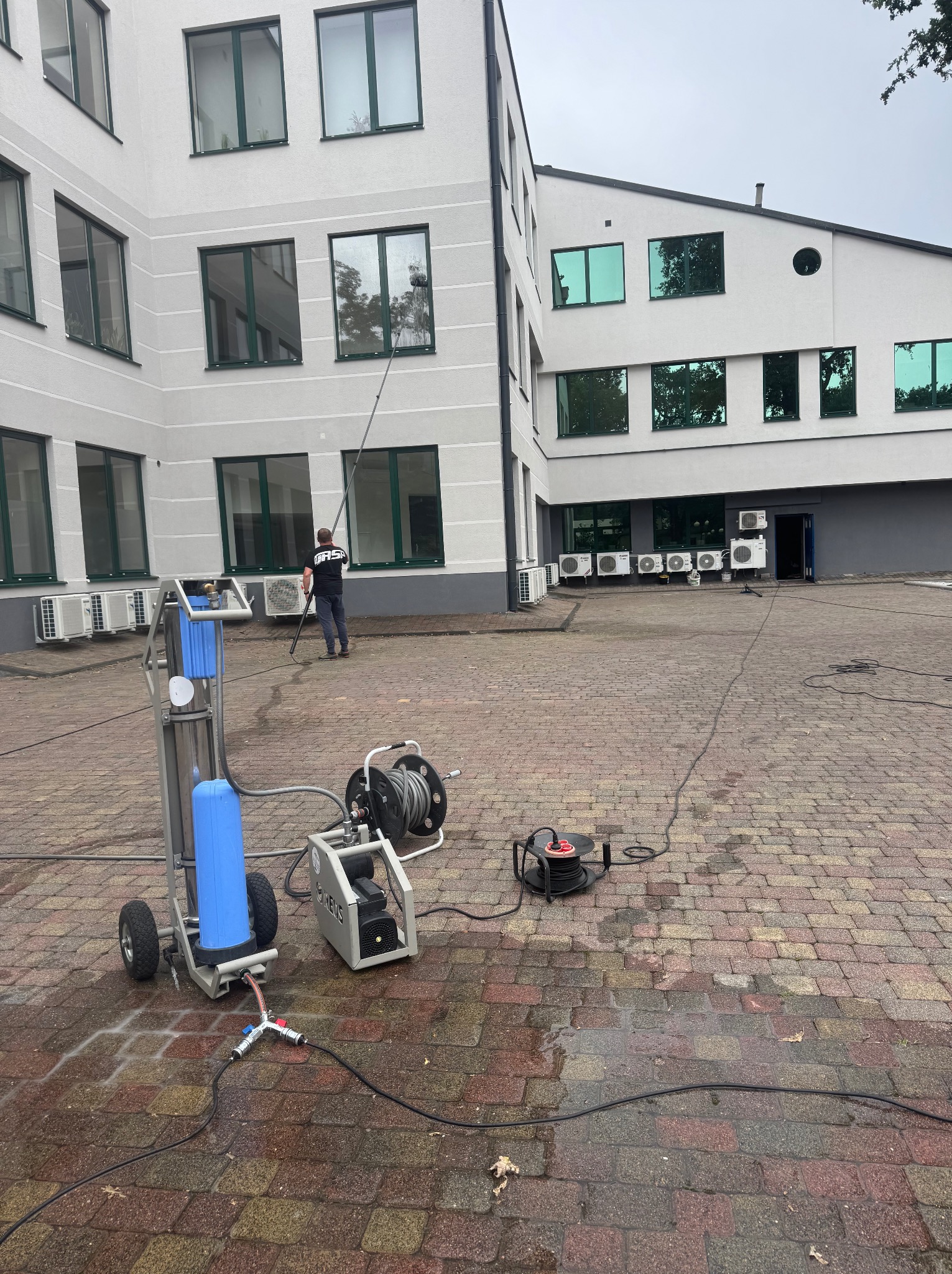 Mycie okien na wysokości przy użyciu specjalistycznego sprzętu. Pracownik w trakcie czyszczenia okien budynku biurowego. Widoczny sprzęt do mycia ciśnieniowego.