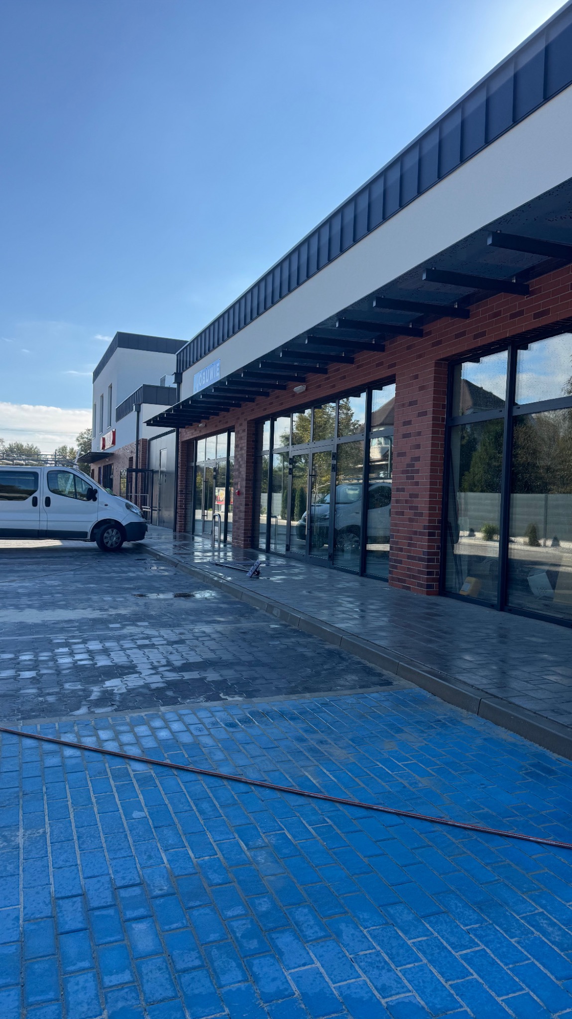 Świeżo umyta elewacja budynku z dużymi oknami i mokrym chodnikiem z kostki brukowej. Biały bus serwisowy zaparkowany obok. Słoneczny dzień.