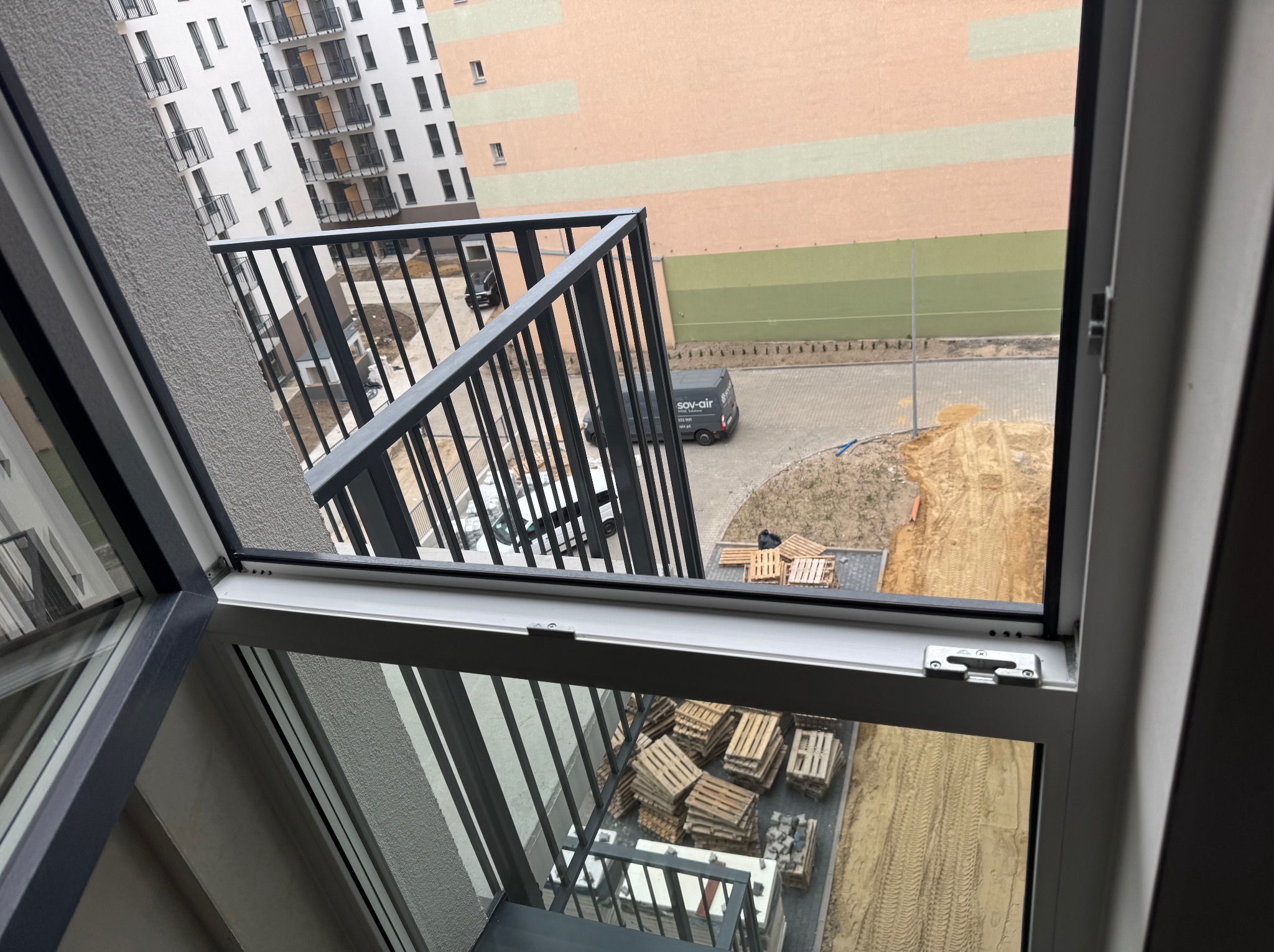 Widok z okna na balkon z czarną balustradą, plac budowy z paletami i budynek. W tle zaparkowany bus firmy Sov-Air. Ujęcie z góry, przez otwarte okno.