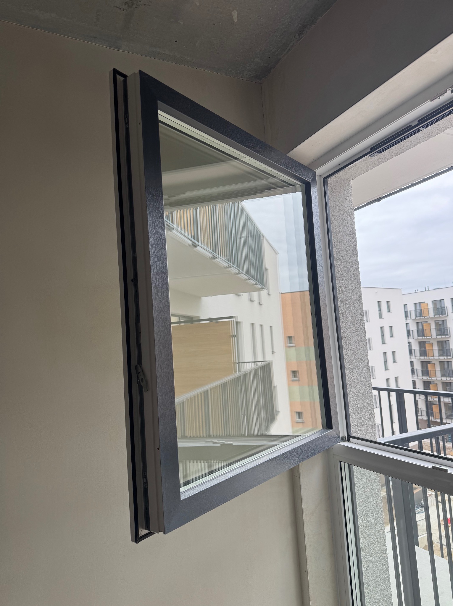 Uchylone okno z szarą ramą na tle balkonu z widokiem na budynki mieszkalne. Widoczny mechanizm otwierania okna.
