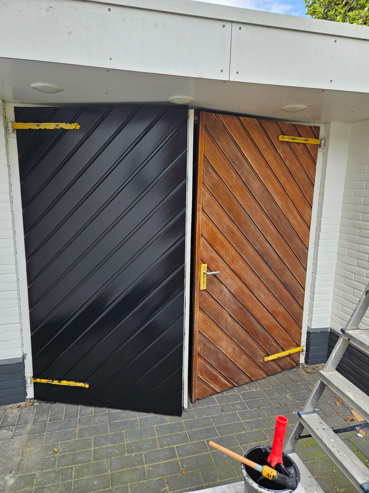 Drewniane drzwi z ukośnymi panelami, jedna część pomalowana na czarno z połyskiem, druga w naturalnym odcieniu drewna. Widoczne akcesoria malarskie i drabina.