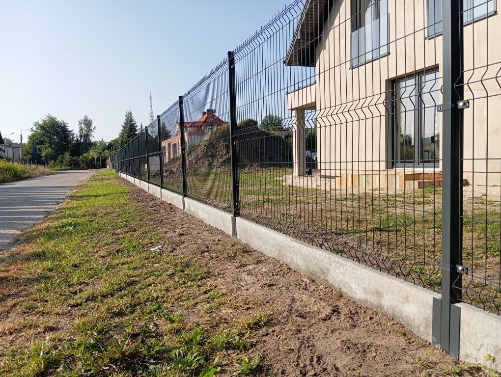 Nowoczesne ogrodzenie panelowe z podmurówką betonową, czarne słupki, widok z zewnątrz posesji, trawnik i dom w tle, słoneczny dzień.