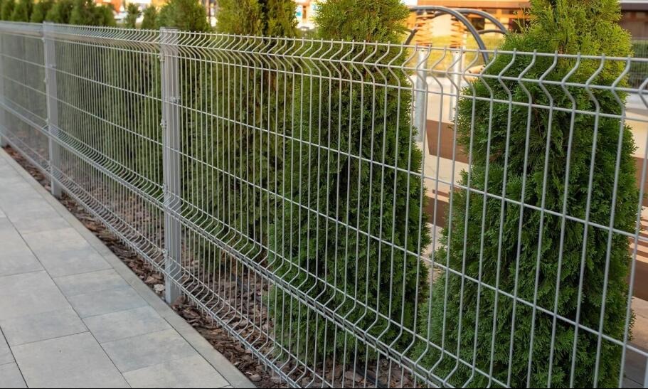 Nowoczesne ogrodzenie panelowe w kolorze srebrnym, z widocznymi, regularnie rozmieszczonymi tujami wzdłuż linii ogrodzenia, na tle kostki brukowej.