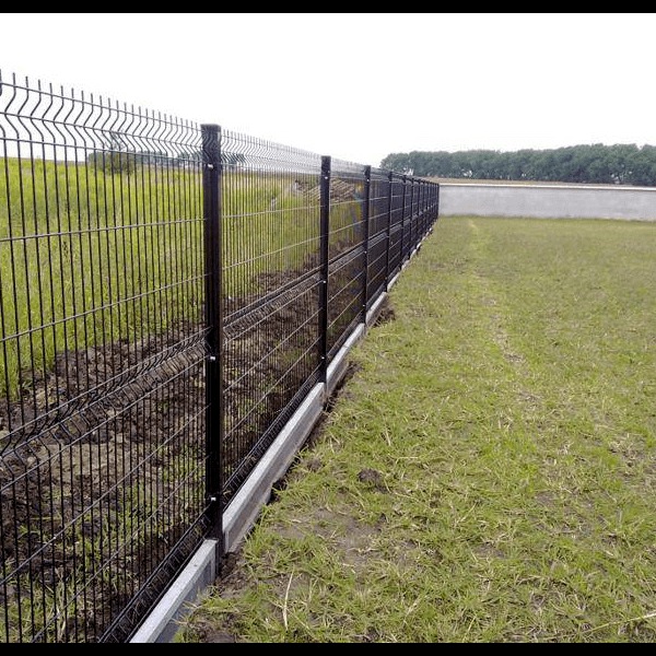 Długie, czarne ogrodzenie panelowe z podmurówką betonową, rozciągające się wzdłuż pola z zieloną trawą i drzewami w tle, widziane z perspektywy bocznej.