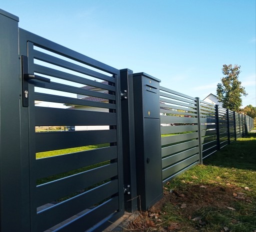 Nowoczesne, poziome przęsła z antracytowej stali tworzą bramę i ogrodzenie. Widoczna skrzynka pocztowa, trawa i drzewo w tle. Lekki kąt z dołu.