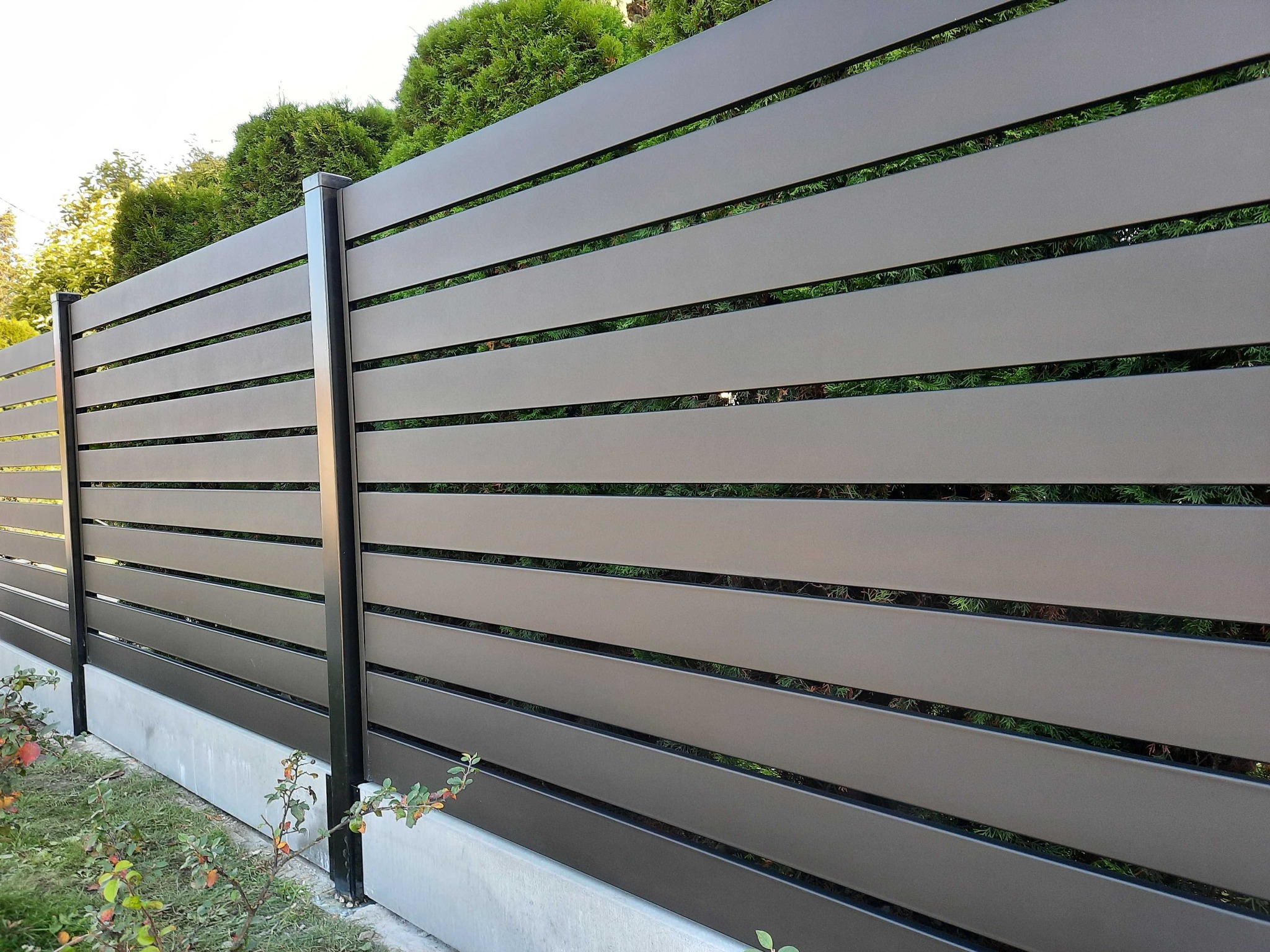 Nowoczesne ogrodzenie panelowe z poziomych, brązowych listew, z czarnymi słupkami, na betonowej podmurówce. Tło stanowią zielone krzewy i trawnik.