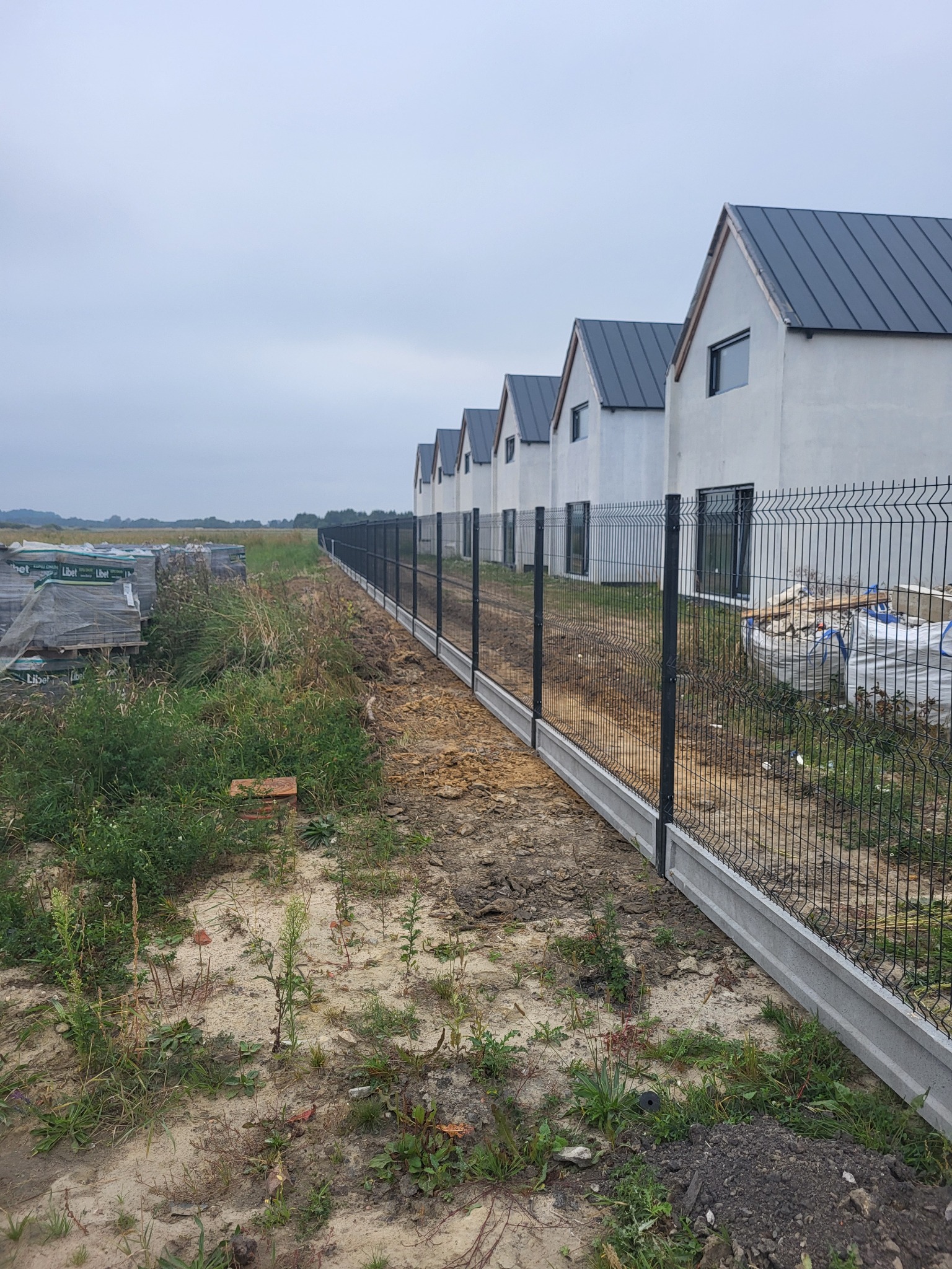 Długa linia nowoczesnego ogrodzenia panelowego z podmurówką betonową, ciągnąca się wzdłuż szeregu domów jednorodzinnych w tle, na tle pochmurnego nieba.