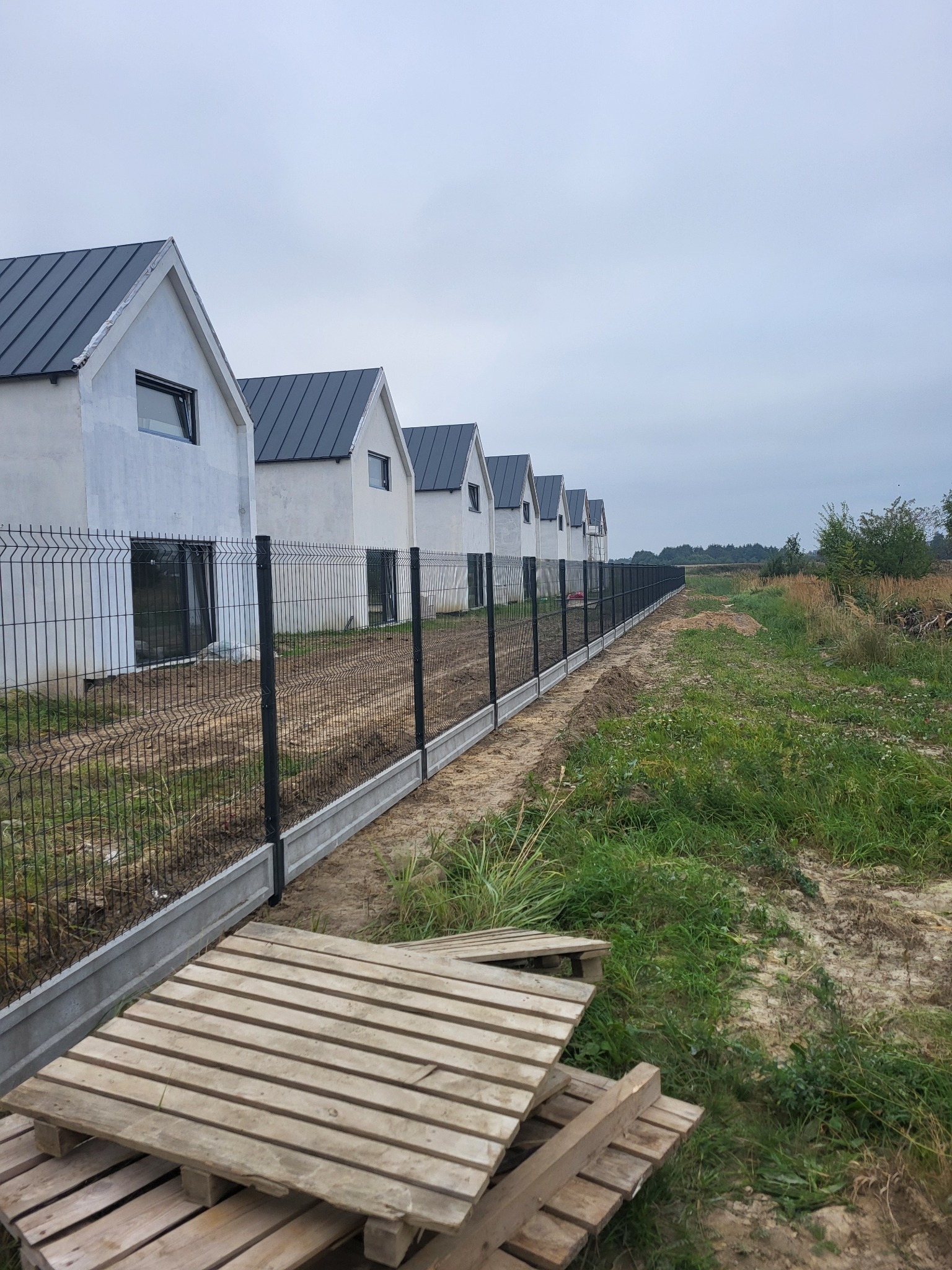 Nowoczesne, czarne ogrodzenie panelowe z podmurówką betonową oddziela szereg identycznych domów od niezagospodarowanego terenu. Na pierwszym planie palety.